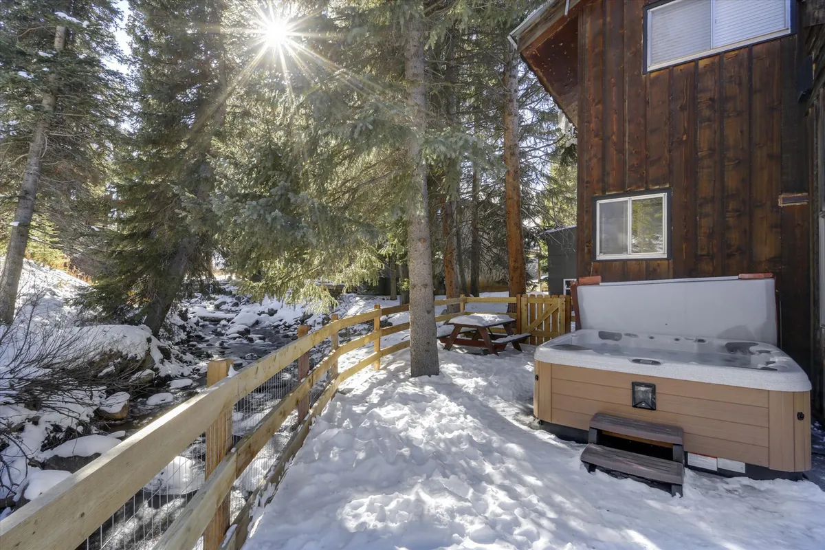 A tranquil riverfront patio scene featuring a bubbling hot tub, a wooden fence bordering the space, a picnic table for gatherings, and a picturesque stream running beside the area. Sunlight filters through the trees, illuminating the scene and highlighting the natural beauty of the surroundings.