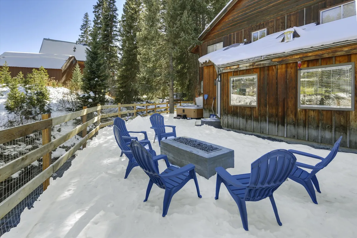 A warm, communal outdoor area with a modern gas fire pit circled by blue chairs, a cozy wooden cabin backdrop, and a snowy forest setting. The scene is lively with hints of recent snow activities and evokes a sense of gathering and play.