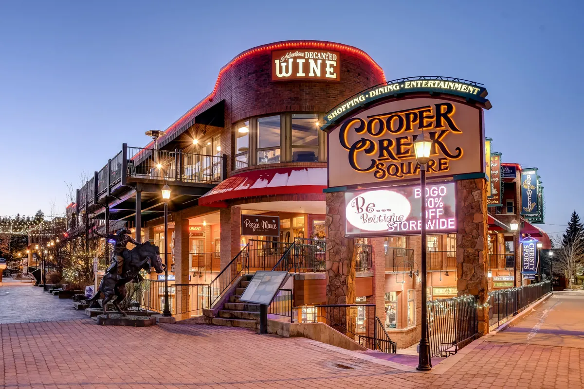 A vibrant scene at Cooper Creek Square, featuring decorative lighting, shopfronts including a wine shop, and people strolling. The area feels lively and welcoming, suggesting opportunities for shopping, dining, and enjoying community events.