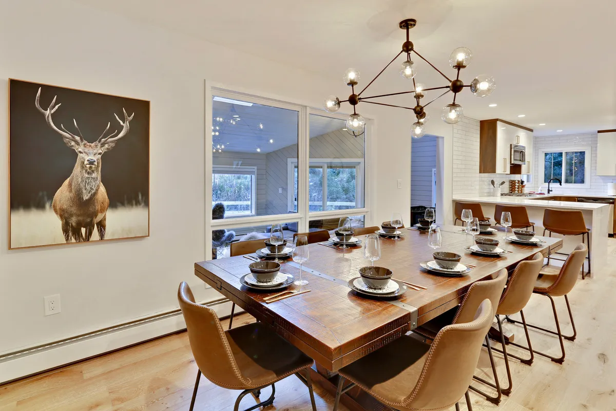 A large dining table set beneath a glowing chandelier, with deer artwork on the wall and windows offering views of the surrounding natural landscape. The setting feels warm and welcoming, designed for family meals and celebrations.