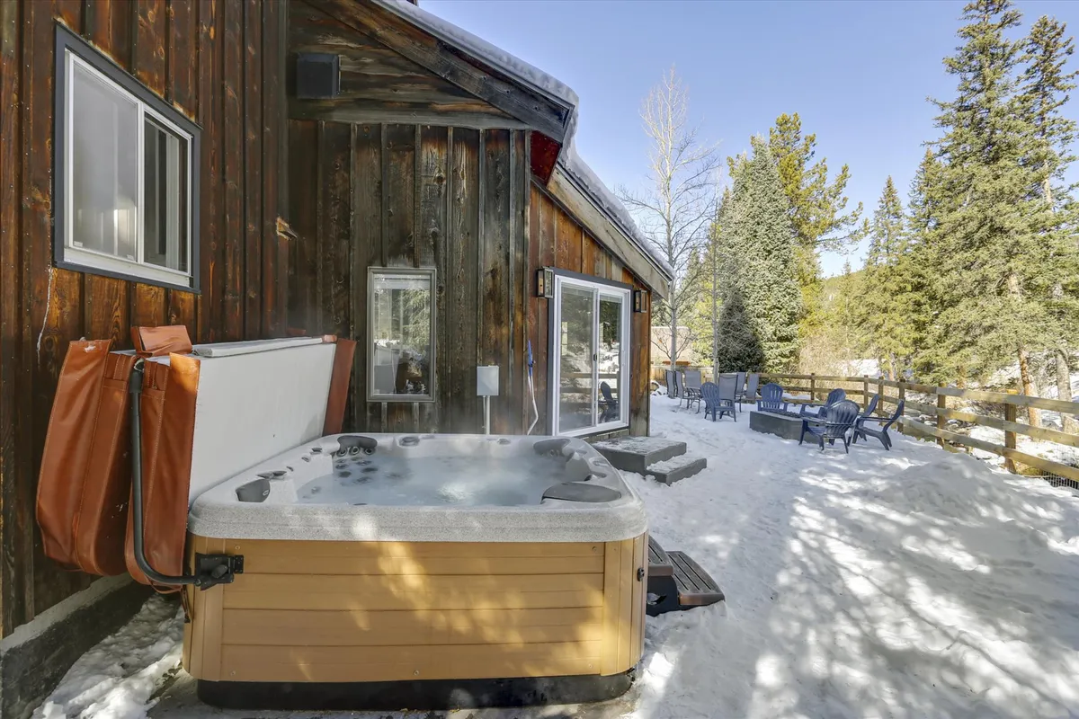 A snow-covered yard frames the inviting hot tub and rustic wooden cabin exterior, surrounded by evergreen trees. The outdoor seating area hints at cozy gatherings with mountain views in a winter wonderland.