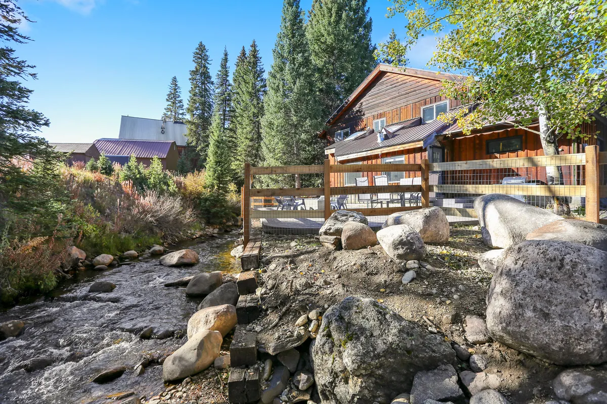 This image captures a wooden cabin nestled beside a flowing creek, surrounded by mature trees and large rocks. The inviting outdoor seating area is arranged to take in the peaceful ambiance of the riverfront location.