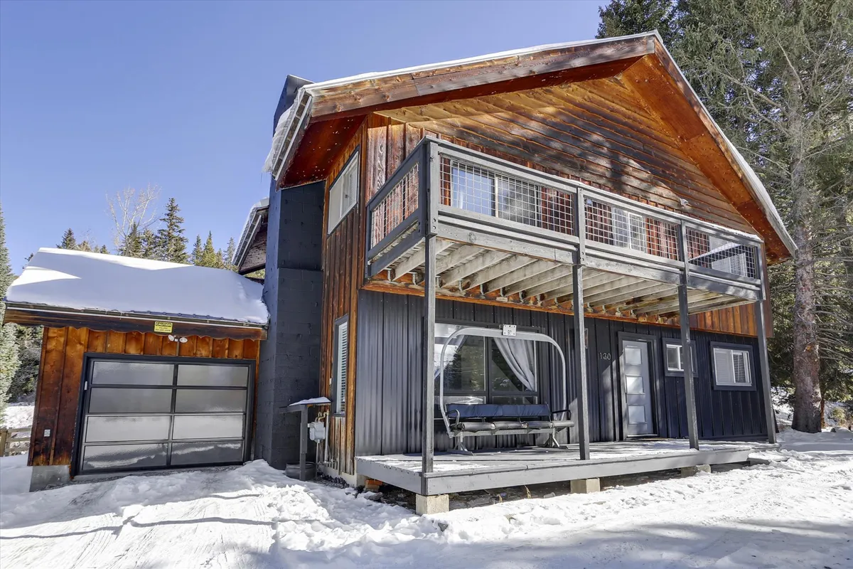 A spacious wooden deck extends from the house, surrounded by snow-covered trees and featuring large windows that blend the indoor and outdoor living spaces. There is ample space for entertaining and enjoying the mountain air, with a modern architectural touch.