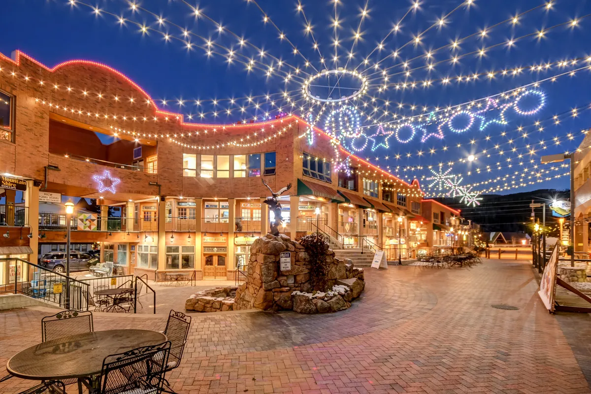 A festive outdoor plaza illuminated by twinkling lights, with a water fountain, decorative stars, and inviting al fresco seating, creating a vibrant community atmosphere.