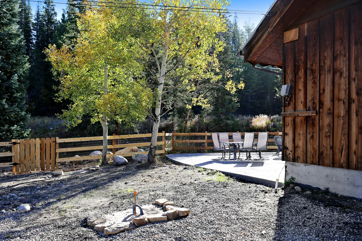 This image showcases a peaceful outdoor seating area with a fire pit surrounded by chairs, set beneath a golden aspen tree in peak autumn color. The area is framed by natural greenery, offering an inviting spot to gather and take in the scenery.