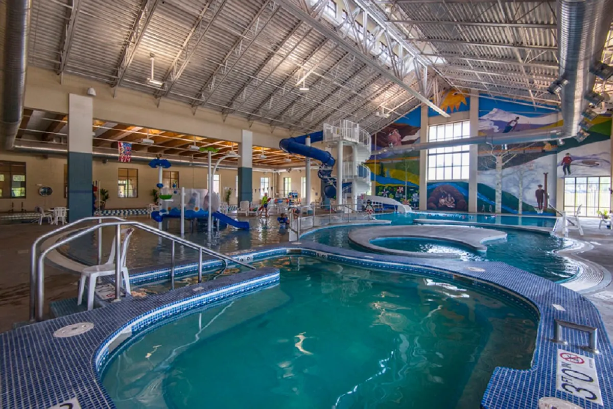 A lively indoor recreational pool with twisting water slides, colorful murals, and lounging areas, filled with families enjoying aquatic activities and play.