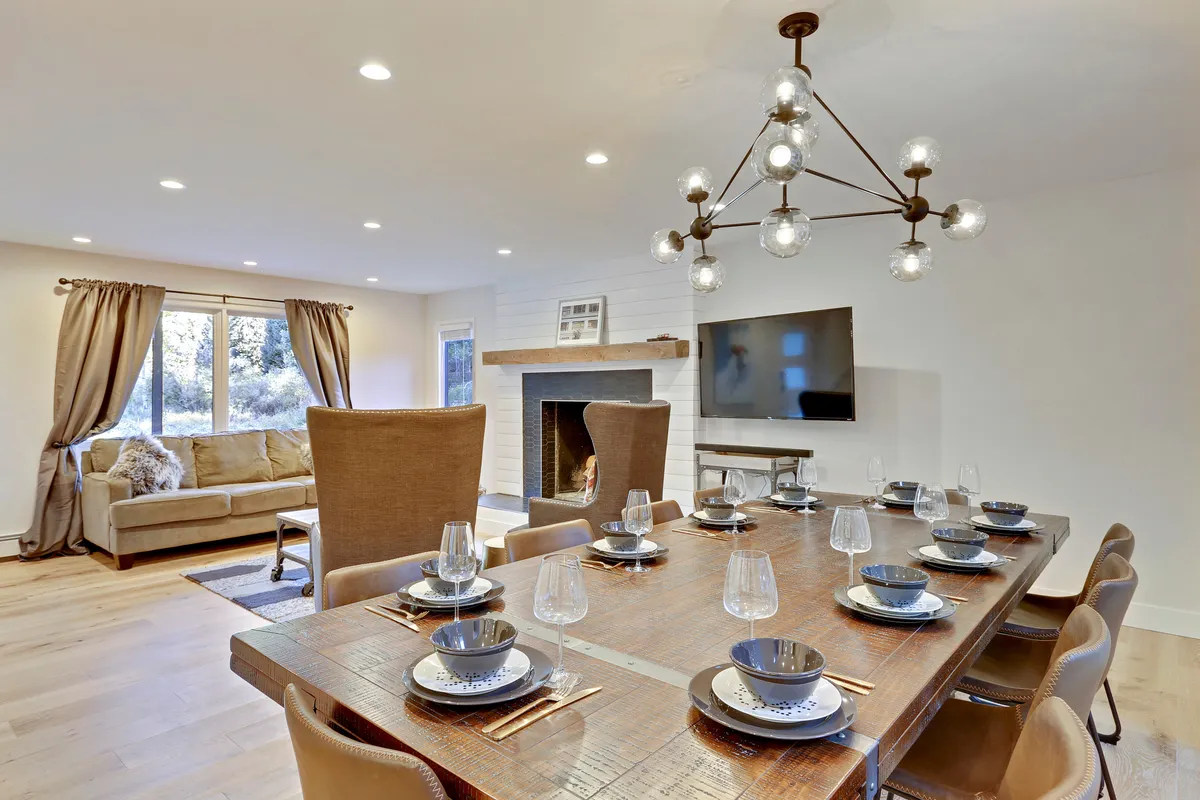 A warm, inviting open-concept living and dining area featuring a large dining table under a modern chandelier, a cozy sofa arranged around a beautiful fireplace, and expansive windows letting in natural light. The space is elegantly styled, seamlessly connecting lounging and dining zones suitable for family gatherings or celebrations.